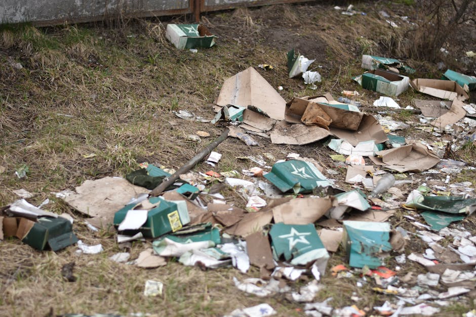 A patch of ground beside a grassy area shows scattered debris, including torn open cardboard boxes with green and white branding, crushed and crumpled along with smaller pieces of paper and packaging 