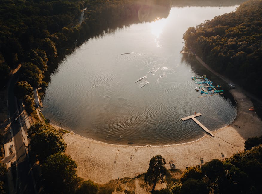 An aerial view of a circular artificial lake surrounded by wooded hills during late afternoon or early evening, with the sun casting a warm glow on the water and trees. The lake's shoreline features a
